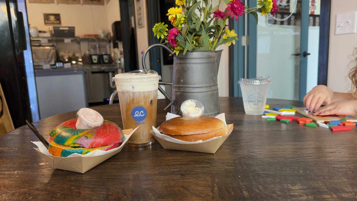 A rainbow raisin bagel, an iced coffee with cream slowly seeping to the bottom, and a plain bagel rest on a round table with an old watering can filled with yellow and pink flowers. A kid’s hands can be seen playing with a puzzle next to a fresh cup of lemonade.
