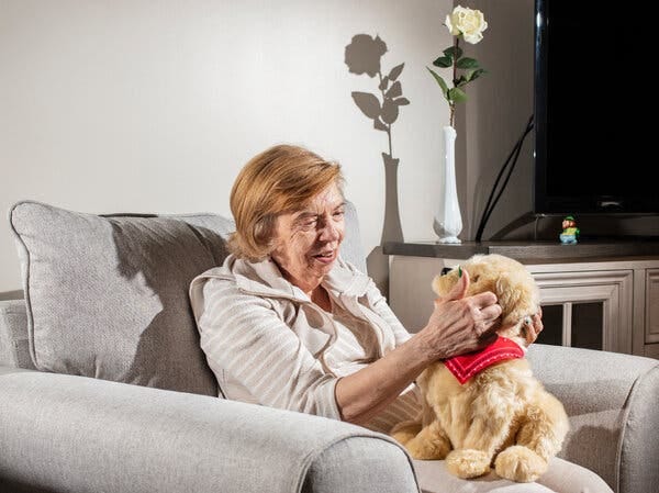 An older woman with red hair sits in a beige armchair caressing a cuddly robot dog. The robot dog, with golden fur, has a red kerchief on its neck and perches on the woman’s lap and looks at her. 
