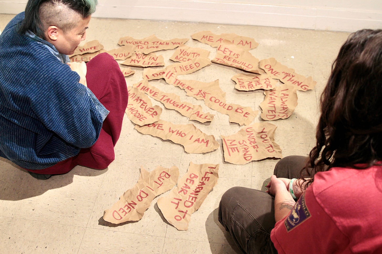 A dozen irregular tan forms, each carrying handwritten text in red, arranged in a loose spread across the concrete floor. Two people sit on either side of the arrangement, looking down at it.