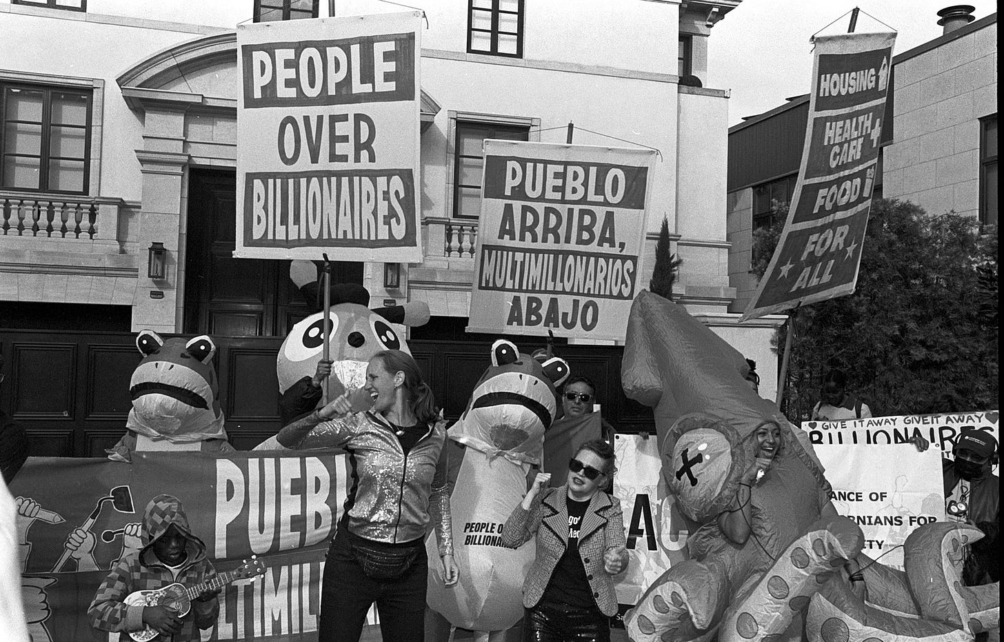 Black-and-white photo of a ‘People Over Billionaires’ protest in San Francisco on Saturday, 11/15. Marchers hold large signs reading ‘People Over Billionaires,’ ‘Pueblo Arriba, Multimillonarios Abajo,’ and banners demanding housing, healthcare, and food for all. Several participants wear inflatable animal costumes during the People Over Billionaires march.