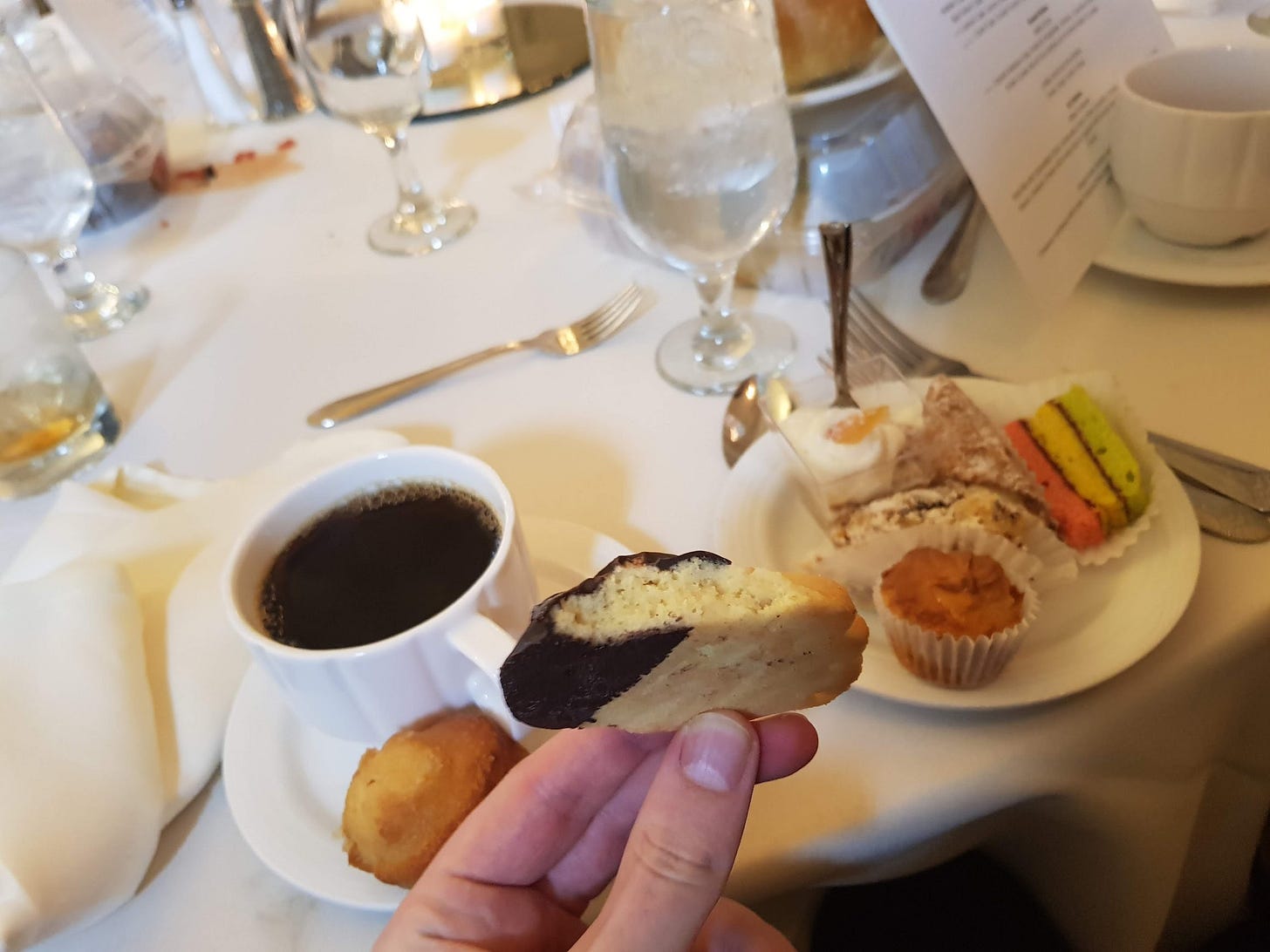 A hand holding a cookie above a fancy table. The table has a cup of coffee and several Italian desserts on it.