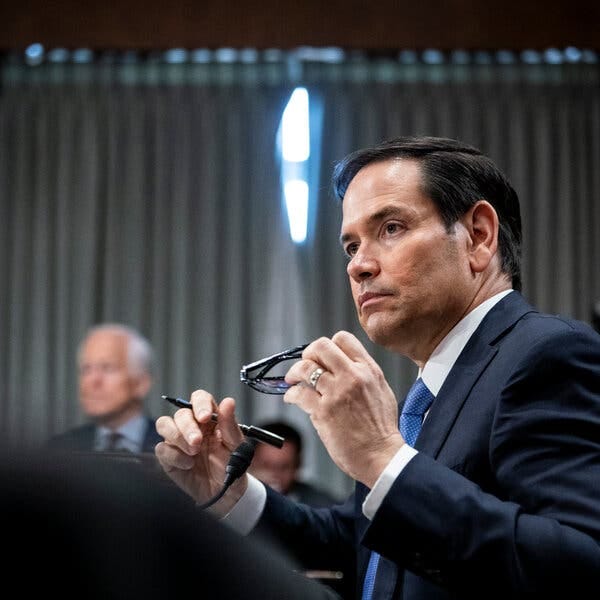 Secretary of State Marco Rubio holds his glasses as he testifies at Capitol Hill. He’s wearing a dark blue suit with a light blue tie.