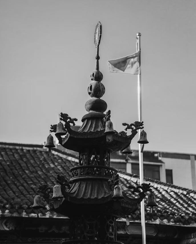 Black-and-white photo of a traditional Chinese temple rooftop ornament adorned with small hanging bells, with a flagpole and partially visible flag in the background.
