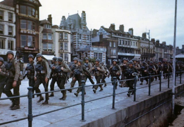 Color image of U.S. troops in combat uniforms marching in two columns at an unspecified port in England, heading to their assigned amphibious shipping. The soldiers look confident and focused.