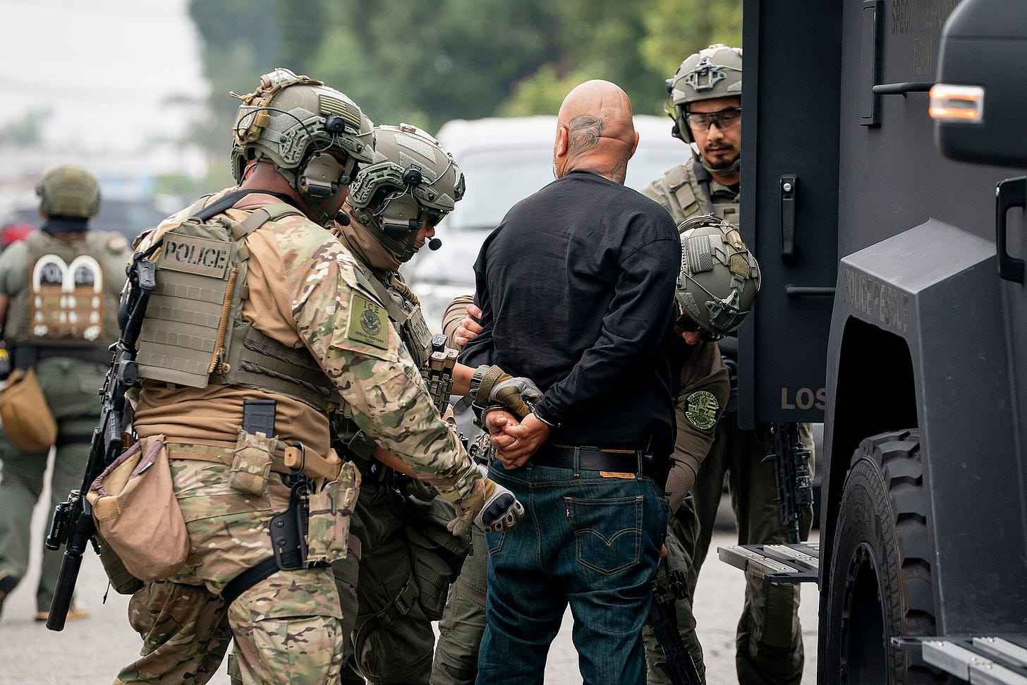 Several ICE agents in full tactical gear arrest a man and handcuff him against a van. His head is turned away.