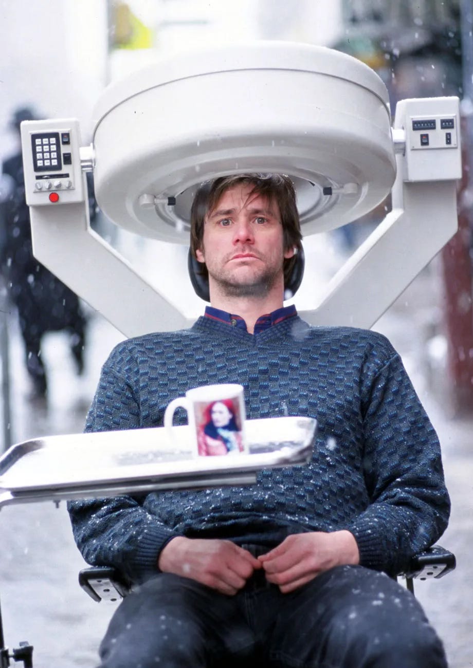 Still from the film Eternal Sunshine of the Spotless Mind. Jim Carrey, a middle aged white man with brown hair, brown eyes, thin lips and a slightly puckered expression, sits in a chair. He’s wearing a raised textured blue sweater, and blue-black pants. It’s snowing and he’s on a sidewalk. On his head is a large white machine with a rounded helmet, V shaped stand, and two panels on opposite sides with buttons. In front of him a mug with the blurry face of the woman he’s trying to forget sits on a metal dentist’s tray.