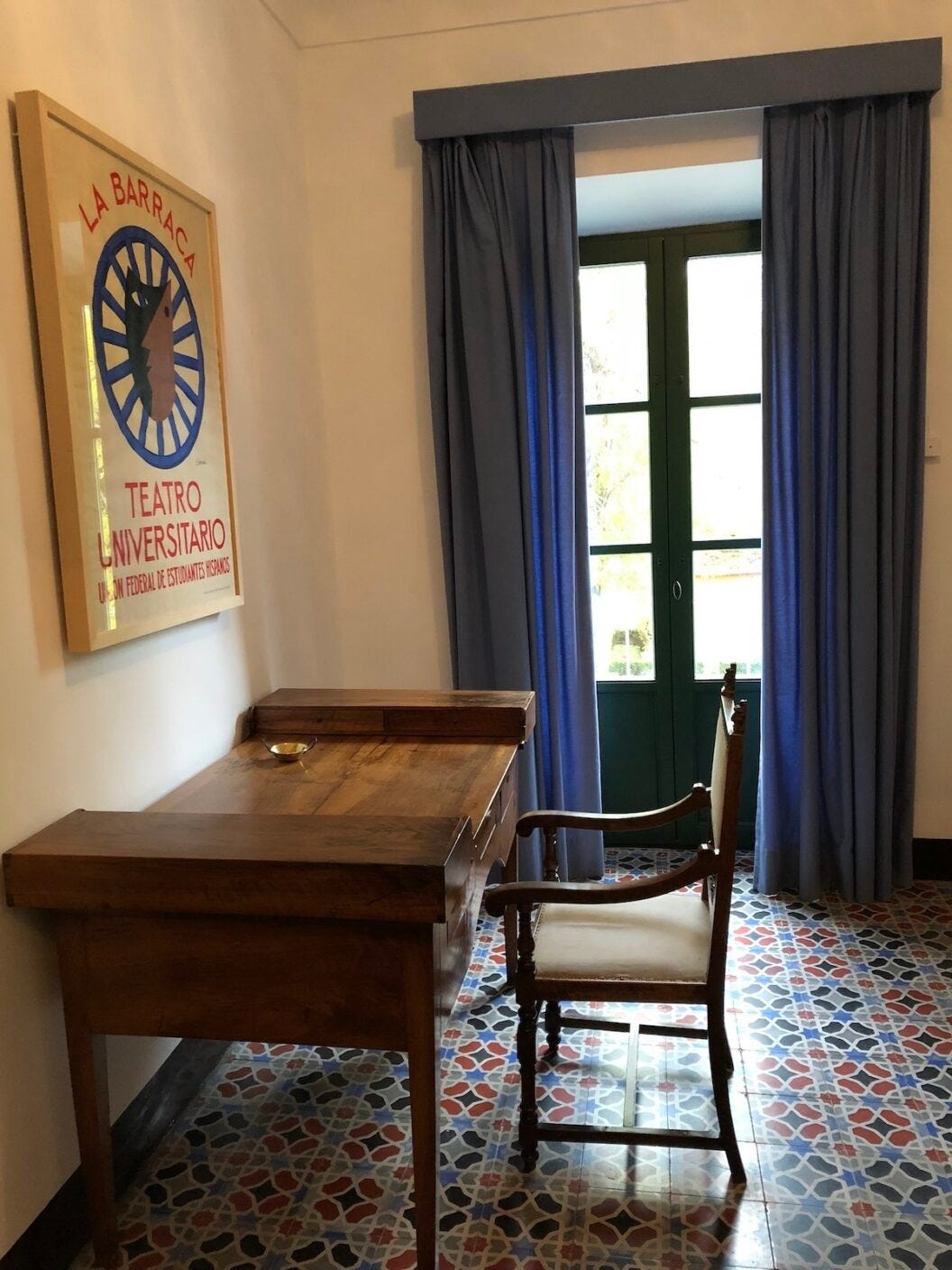 1200 A wooden desk and chair sit in a room with colorful mosaic tile flooring, blue curtains, and a framed "La Barraca Teatro Universitario" poster on the wall.