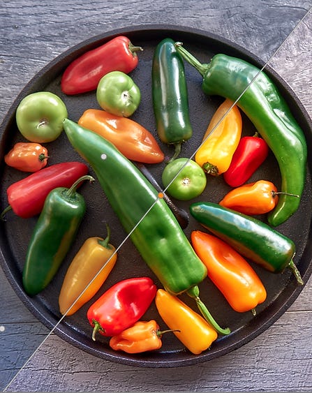 Comparison of Unedited vs Edited View of Photo  Unedited View: The unedited photo presents a vibrant assortment of fresh peppers and green tomatoes arranged on a dark, round tray. The colors are rich and varied, showcasing the natural beauty of the garden produce. However, the background may appear slightly dull or less defined, which can detract from the overall impact of the image.  Edited View:  In the edited version, enhancements are made to amplify the vibrancy of the colors and improve the contrast between the produce and the textured background. The peppers and tomatoes appear more vivid, drawing the viewer's attention. Additionally, any distractions in the background may be minimized, allowing the focus to remain on the fresh produce, creating a more appealing and polished final image.