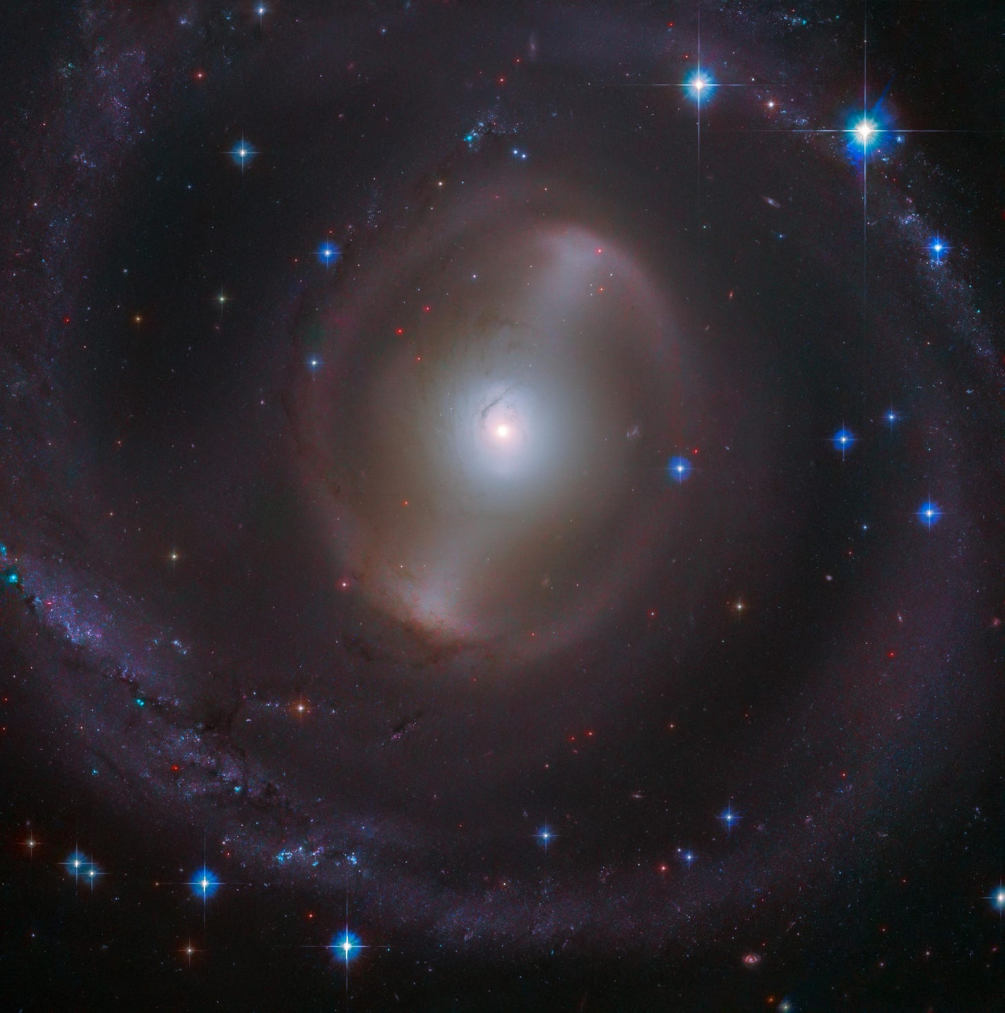 A bright, white galactic core shines near the center of the image with a faint bar of stars extending from it, diagonally to the right. Faint, hazy spiral arms encircle the core, with several distant stars and bright blue foreground stars with diffraction spikes scattered throughout the image, all against black space.