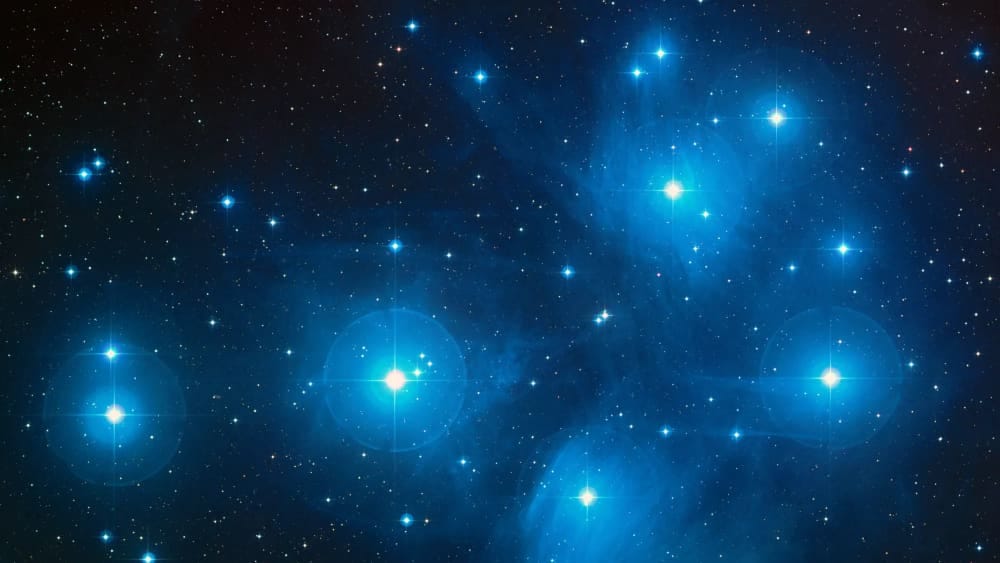A photo of the Pleiades star cluster depicting an array of blue-white stars shimmering in space A photo of the Pleiades star cluster depicting an array of blue-white stars shimmering in space