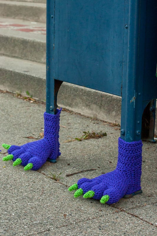 Close-up of a blue Post Office mail box with hand knitted blue booties with green talons made to look like furry monster's feet.