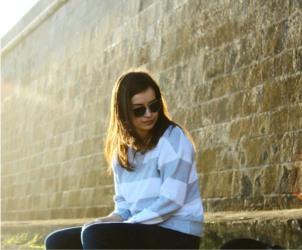 Young woman with dark brown hair and sunglasses is sitting on a stone bench that is part of a high stone wall. She is looking down to her left, studying an orange hued orb that appears to be sitting close by on the bench with her. There is also a slightly orange hue behind the left side of her hair. Young woman with dark brown hair and sunglasses is sitting on a stone bench that is part of a high stone wall. She is looking down to her left, studying an orange hued orb that appears to be sitting close by on the bench with her. There is also a slightly orange hue behind the left side of her hair.