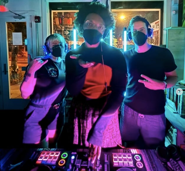 Trois personnes portant un masque noir FFP2 posent devant une table de mixage éclairés par une lumière rose