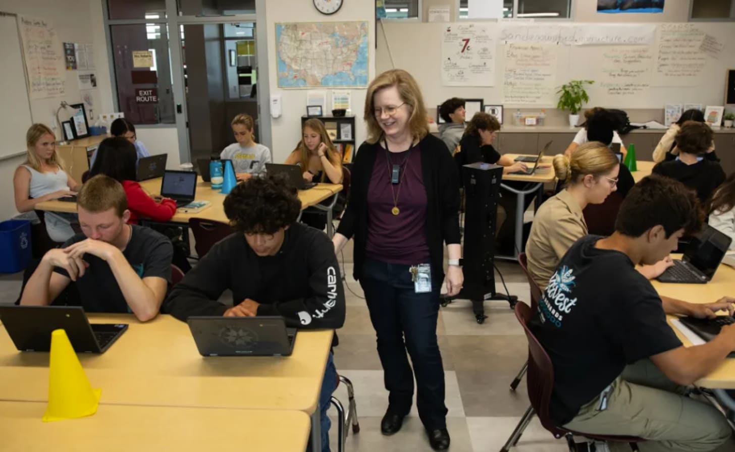 A teacher walking between rows a students on devices, each of them looking quite bored.