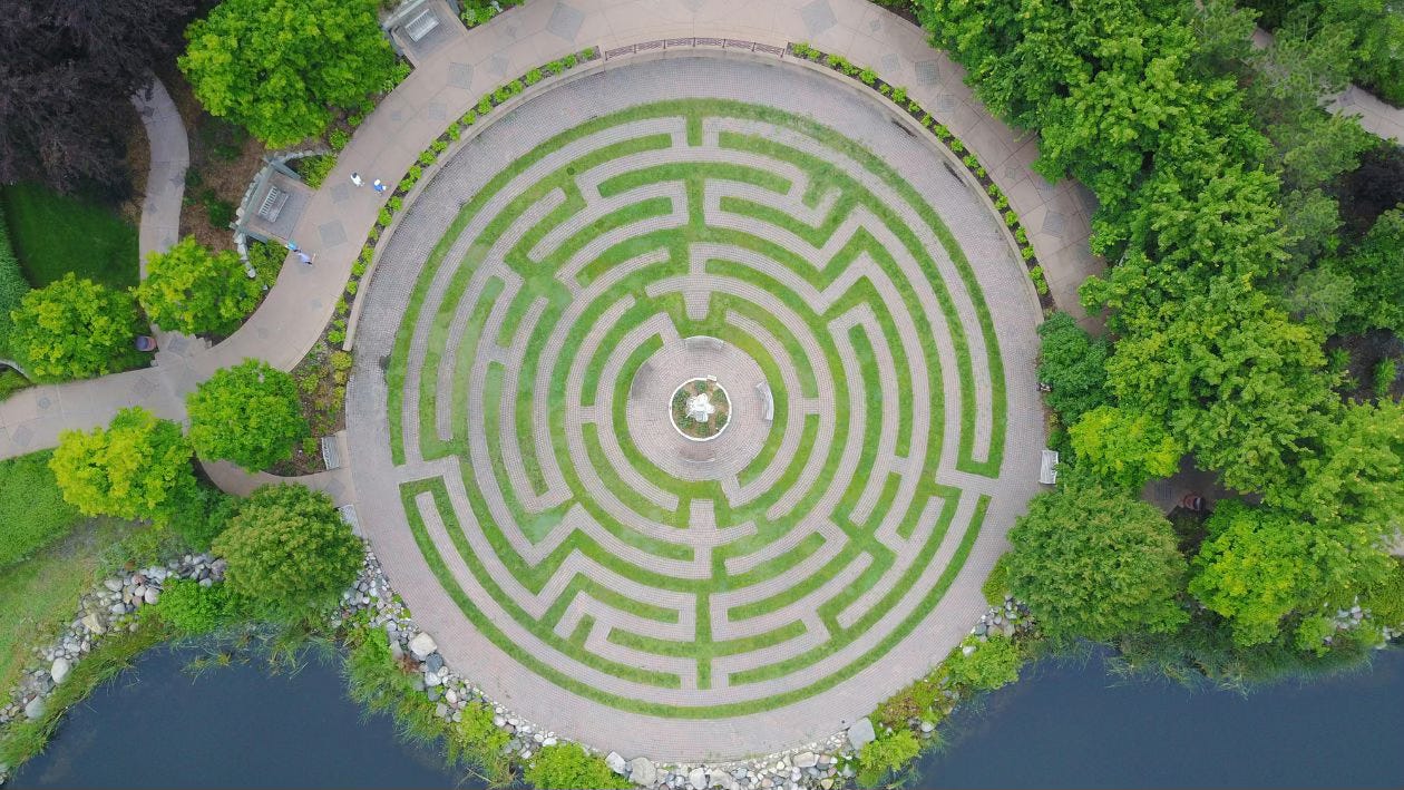 Aerial shot of a spiraling labyrinth of green hedgerows, with a central gathering spot at its heart. Forest and deep waters surround it.