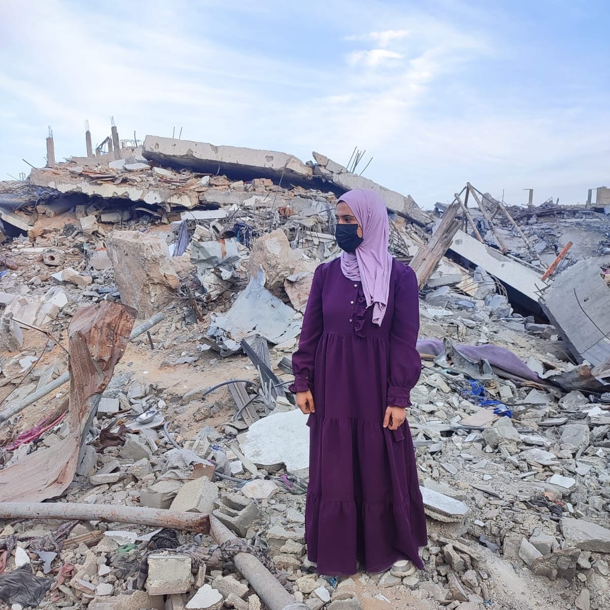 A woman wearing a long purple dress, a lavender hijab, and a black face mask stands amid the rubble of collapsed concrete buildings. The background shows extensive destruction, with broken slabs, twisted rebar, and debris scattered across the ground under a partly cloudy sky.