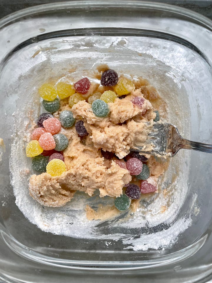 A bowl of butter and sugar with red ketchup swirling in, and a bowl of cookie dough with a pile of multicolored gumdrops stirring in