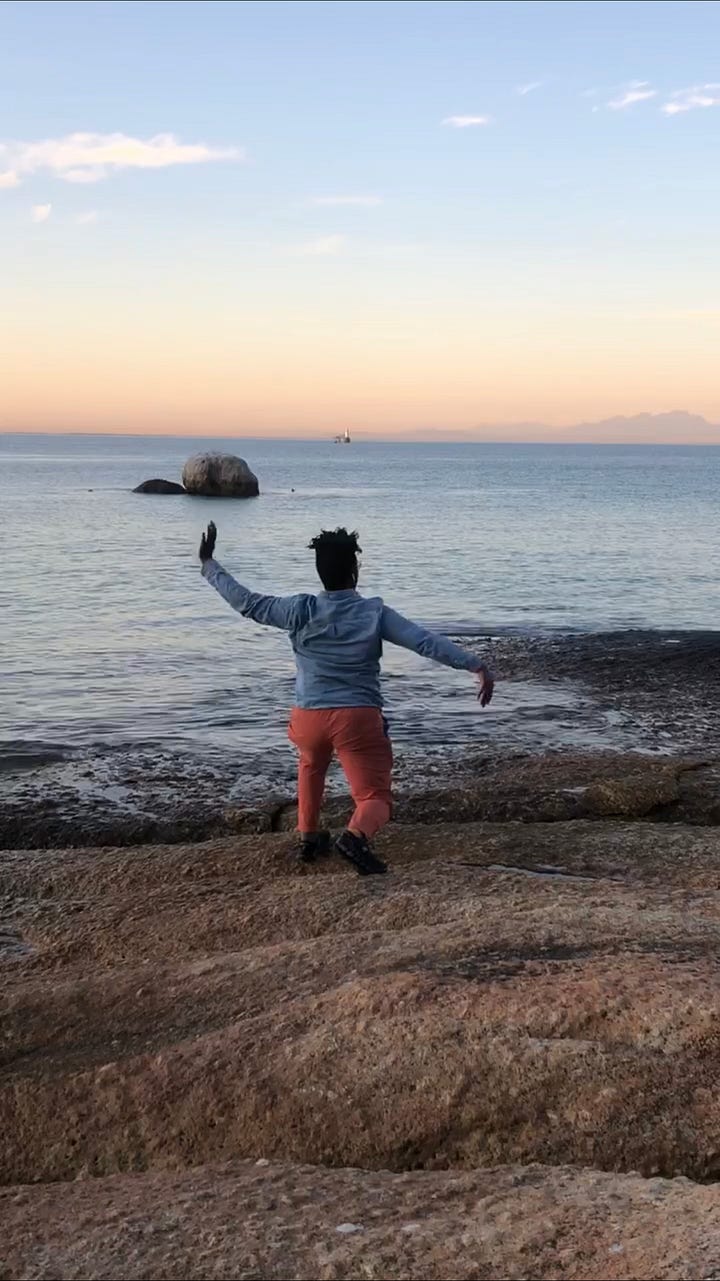 Two images of me moving across the ocean in Cape Town, South Africa. I'm wearing orange pants and a blue shirt. In the distance is an orange sky, the mountain line, boulders and faint blue water.