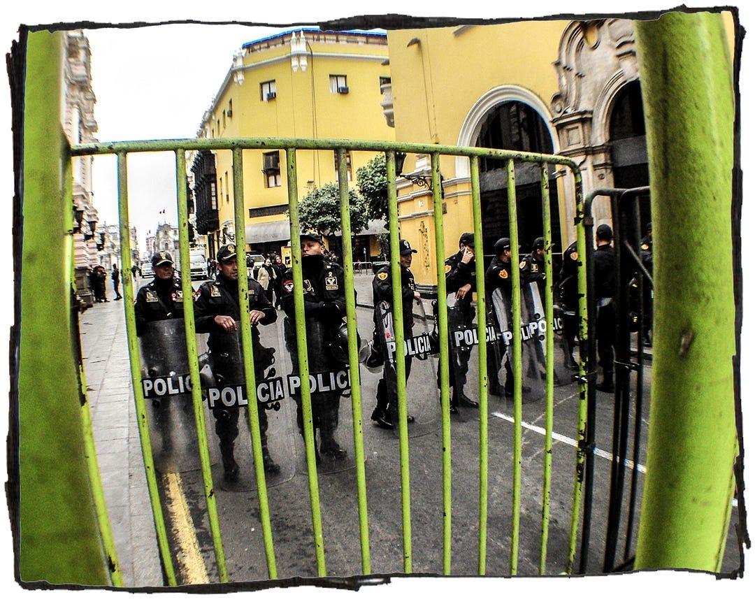 An image of a group of police officers with riot shields standing behind a green metal fence.