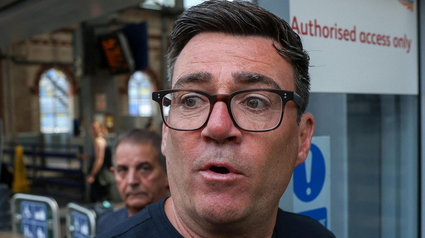 Andy Burnham pictured in the centre of the frame, wearing dark-framed glasses and a dark navy or black top. The photograph is tightly cropped around the head and shoulders. His face is sharply in focus, while most of the background is blurred. Behind him is a railway station concourse. A second man is visible in the background, but out of focus.