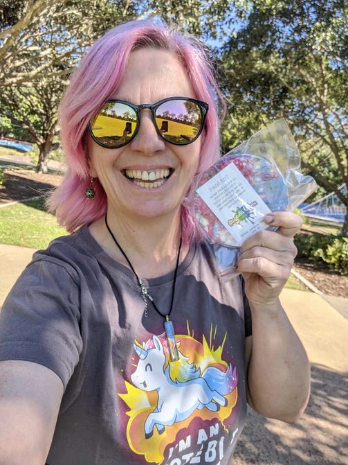 A smiling woman with pink hair and sunglasses stands in a sunny park, holding a round piece of found art wrapped in clear plastic. She wears a dark t-shirt with a cartoon unicorn and the text “I’m an out of this world zombie.” The artwork label reads “Found Art – Free Gift” and includes Magenta Quinn’s website. A small round artwork wrapped in clear plastic sits on a stone surface overlooking a coastline. Trees sway in the background with the ocean visible beyond, creating a peaceful setting for the surprise art drop. A label on the package invites passersby to take the piece as a gift.