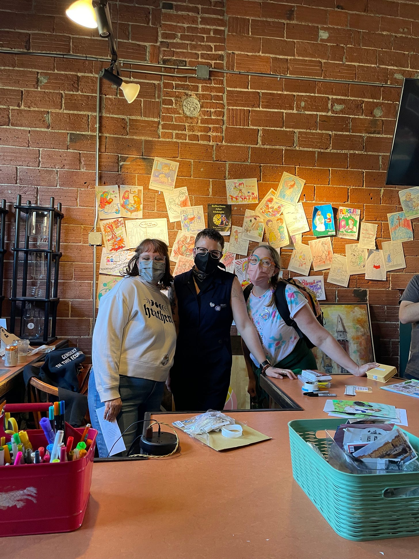 three white humans wearing masks stand in front of a brick wall with dozens and dozens of coloring pages taped up to a brick wall behind them