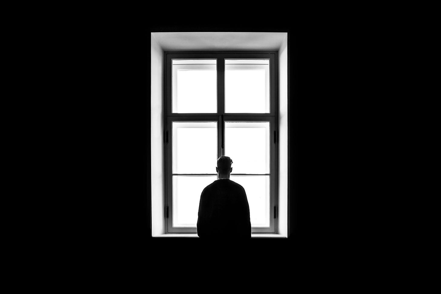 A silhouette of a person standing in a dark room,looking out a window. The window is illuminated in white and has six panes. A silhouette of a person standing in a dark room,looking out a window. The window is illuminated in white and has six panes.