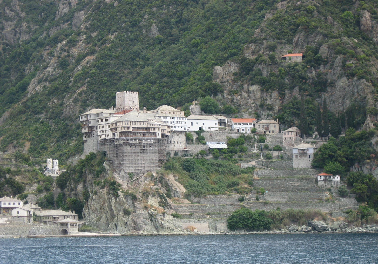 Multistory stone and white-plastered monastery buildings with a fortified tower, built on a cliff above the sea, backed by a wooded mountainside.