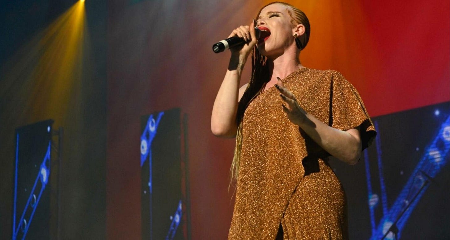 A femme singer with albinism is singing into a mic on a large stage. She wears a regal gold dress, red lipstick, and braids.