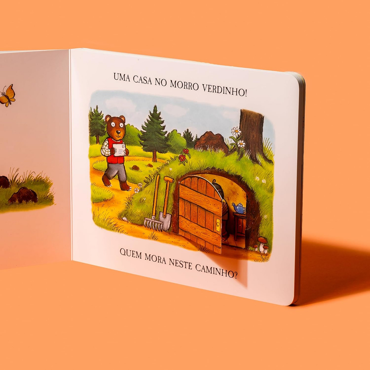 #descrevemospravocê  A imagem mostra um livro infantil aberto, posicionado sobre uma superfície lisa na cor laranja, com fundo da mesma cor. A página visível apresenta uma ilustração colorida e alegre. Na parte superior da página, está escrito: Uma casa no morro verdinho! e, na parte inferior, a pergunta: Quem mora neste caminho?  A ilustração mostra uma cena campestre, com um morro coberto de grama verde, árvores ao fundo e um céu azul claro com nuvens. No centro da imagem, há uma porta de madeira aberta embutida no morro, revelando o interior aconchegante de uma casa subterrânea, onde se vê parte de uma cômoda com um bule azul em cima. Do lado de fora, encostadas na grama ao lado da porta, estão ferramentas de jardinagem, como uma pá e um ancinho. À esquerda da cena, um urso marrom em pé caminha em direção à casa, sorrindo e segurando uma caneca. Ele veste camisa branca, colete vermelho e calça cinza. Há detalhes florais pelo caminho e, à esquerda da dobra do livro, aparece parcialmente a imagem de uma borboleta voando sobre a grama.  A composição transmite uma atmosfera acolhedora e curiosa, típica de livros para primeira infância, com traços simples e cores vibrantes.
