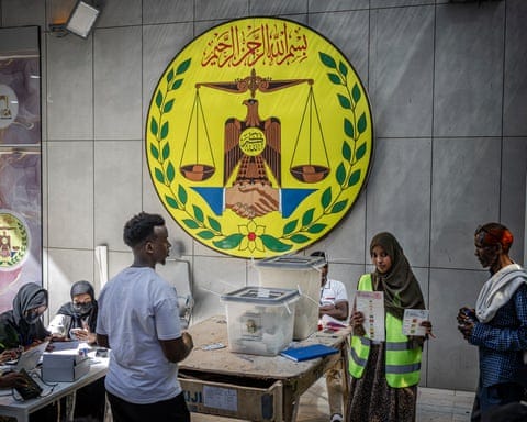 People work in a polling station with a plastic ballot box on a table and the national emblem of Somaliland on a wall