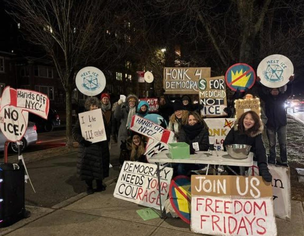 Nighttime photo of about a dozen people in winter gear on the pedestrian path of a parkway, with Freedom Friday signs ("Honk 4 Democracy," "Melt Ice," "Democracy Needs Your Courage," "Join Us Freedom Fridays," "Hands Off NYC! No ICE.") Two people are seated behind a table with 2 large bowls on it for whistle and zine distribution. Nighttime photo of about a dozen people in winter gear on the pedestrian path of a parkway, with Freedom Friday signs ("Honk 4 Democracy," "Melt Ice," "Democracy Needs Your Courage," "Join Us Freedom Fridays," "Hands Off NYC! No ICE.") Two people are seated behind a table with 2 large bowls on it for whistle and zine distribution.