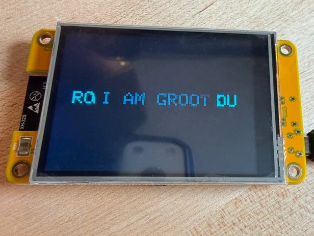 Photo of a yellow ESP32 display on a wooden surface showing blue pixel text that reads “I AM GROOT” with partial letters on a dark screen.