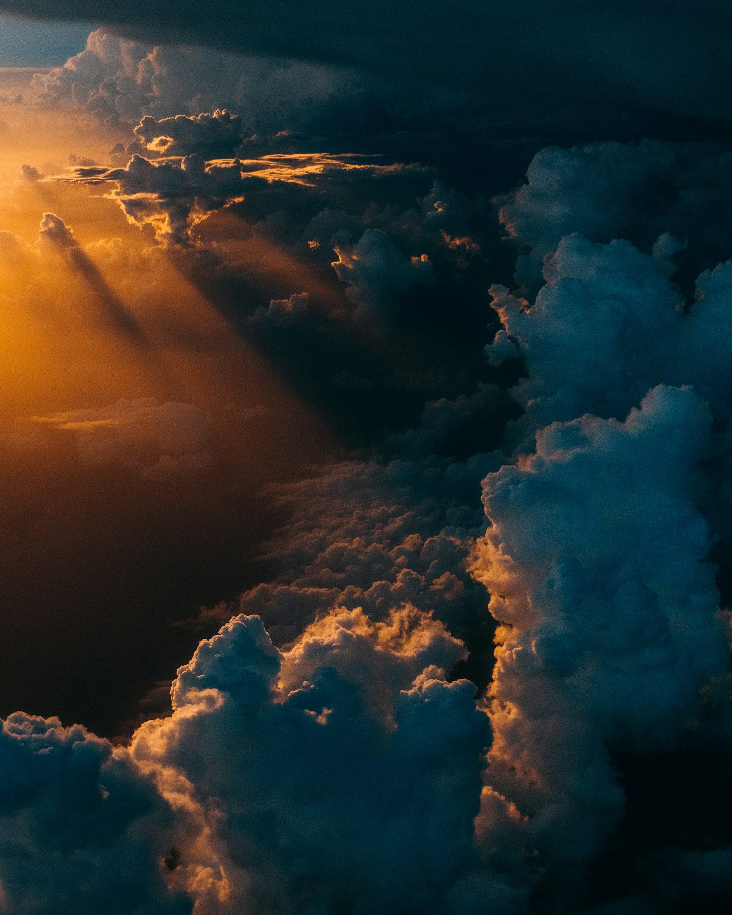 low cumulus clouds, grey backed against the light, rise on the right to storm-like column and bands of higher clouds in the distance, all offset by golden-orange beams of light coming from the upper left