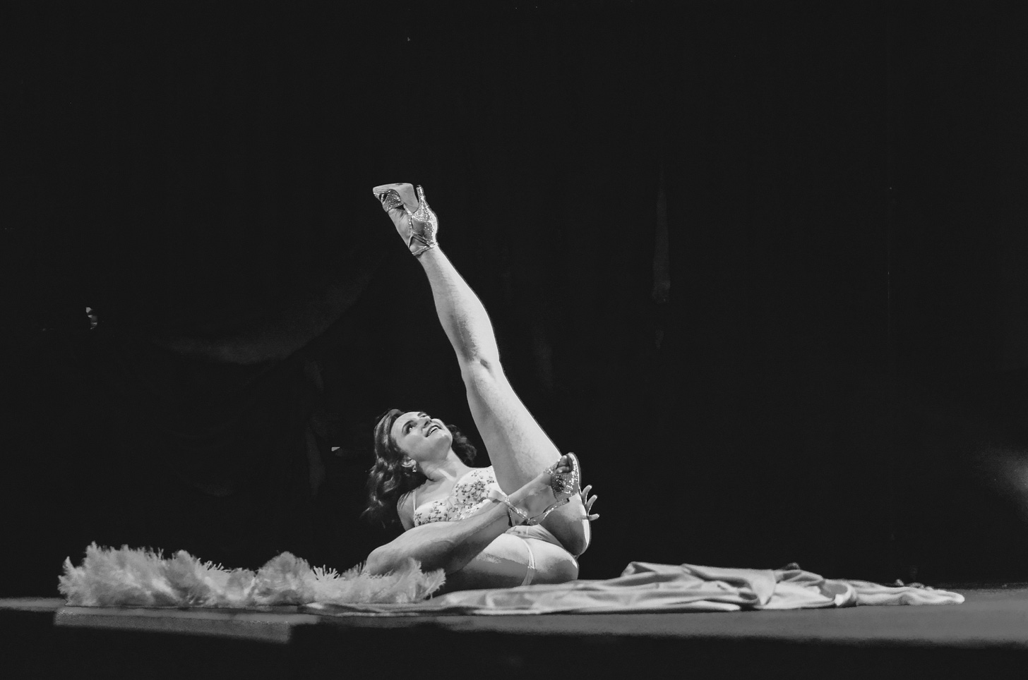A black and white photo of Halena on stage performing a burlesque solo. Halena is on the ground with one leg in the air, focusing only on that one leg. She is wearing white and there are feather fans on the stage beside her.