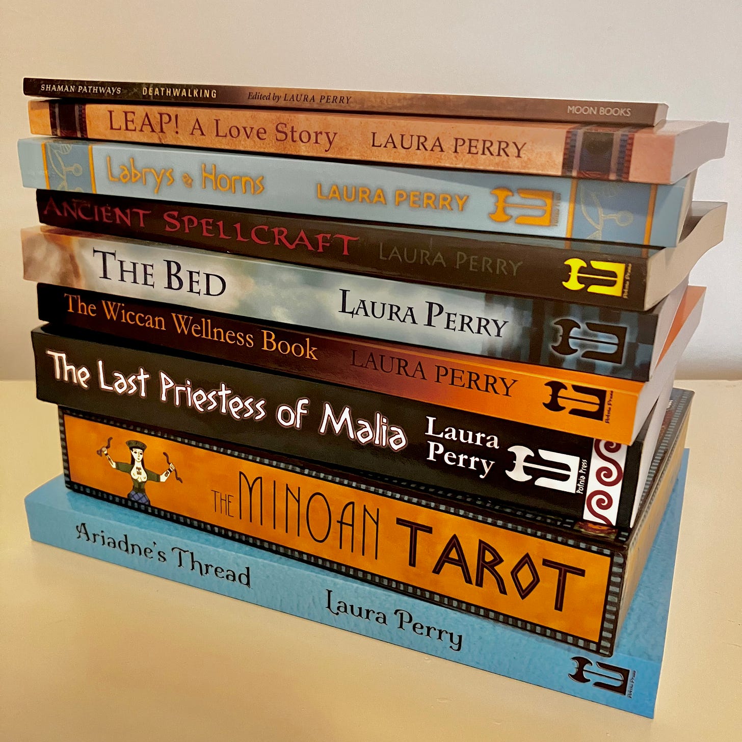 A stack of books Laura has written, with the spines facing the camera. From the bottom, the titles read Ariadne's Thread, The Minoan Tarot, The Last Priestess of Malia, The Wiccan Wellness Book, The Bed, Ancient Spellcraft, Labrys & Horns, Leap! A Love Story, and Deathwalking.