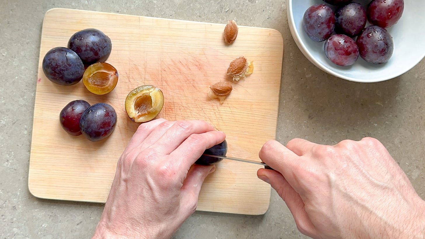 Oblong purple plums on a cutting board, while Martin slices them in half and removes the pits.