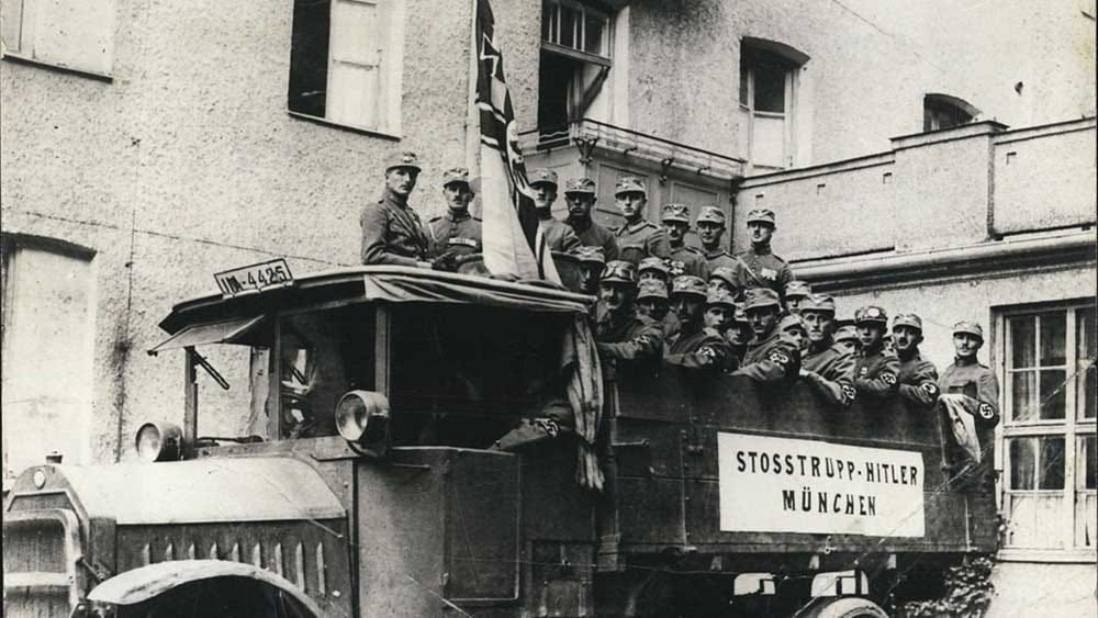 Historical black and white photograph showing uniformed men of the Stoßtrupp-Hitler standing on and around a truck in Munich in 1923, with a sign identifying the unit mounted on the vehicle's side.