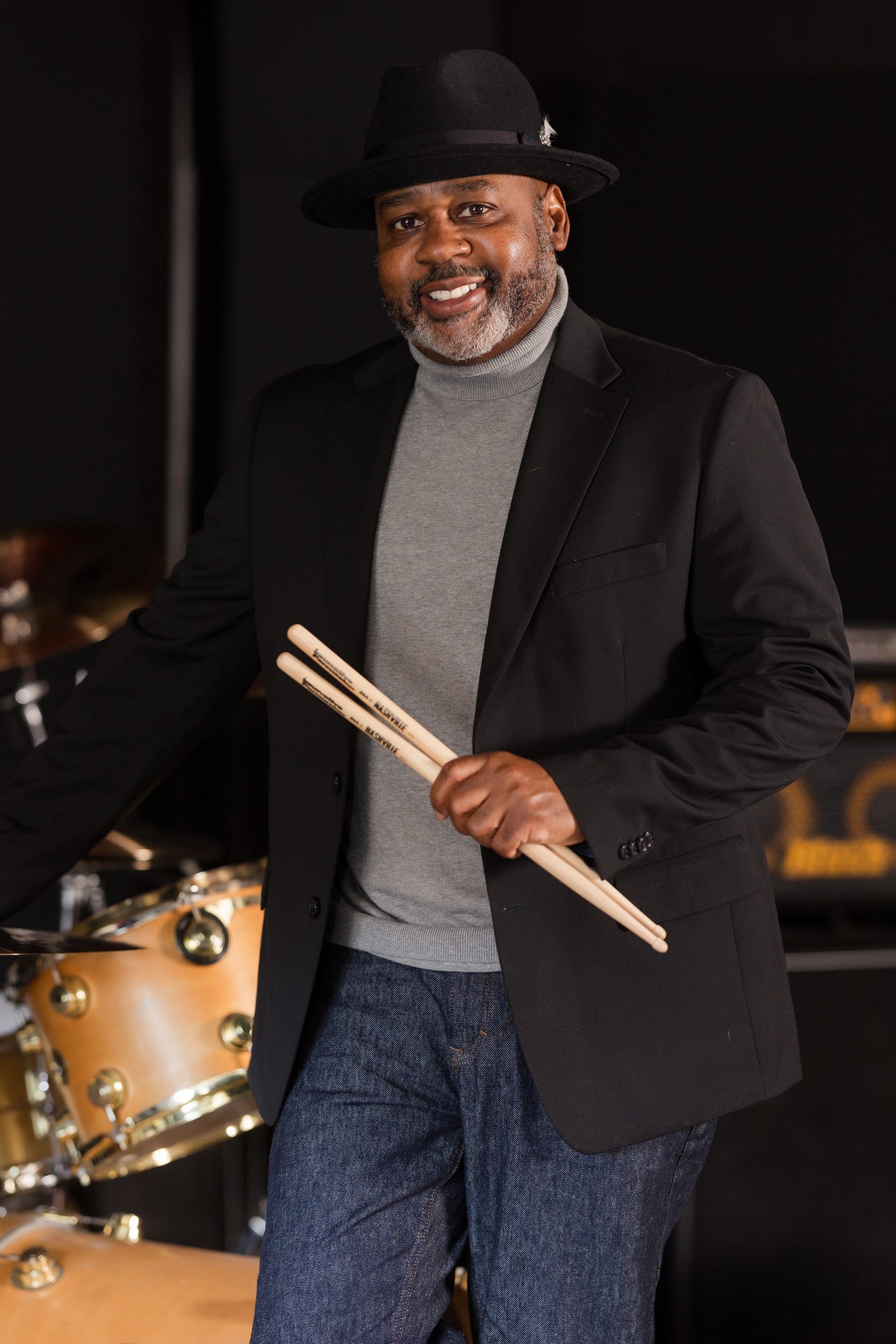 Clayton Craddock smiling in a black hat and holding drumsticks