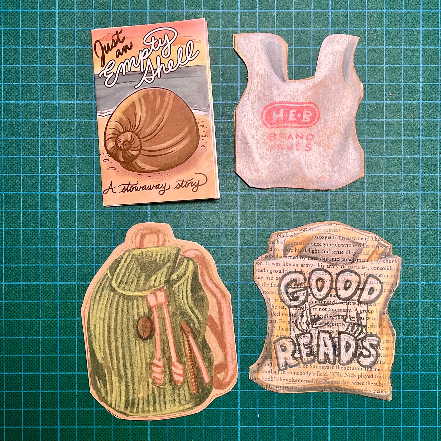 Photo des 4 mini zines posés sur mon tapis de découpe : le premier a un dessin de coquillage en couverture, le deuxième est en forme de sac de supermarché de la marque HEB, le troisième est en forme de sac à dos et le quatrième est découpé en forme de sac en papier de la marque Good Reads.