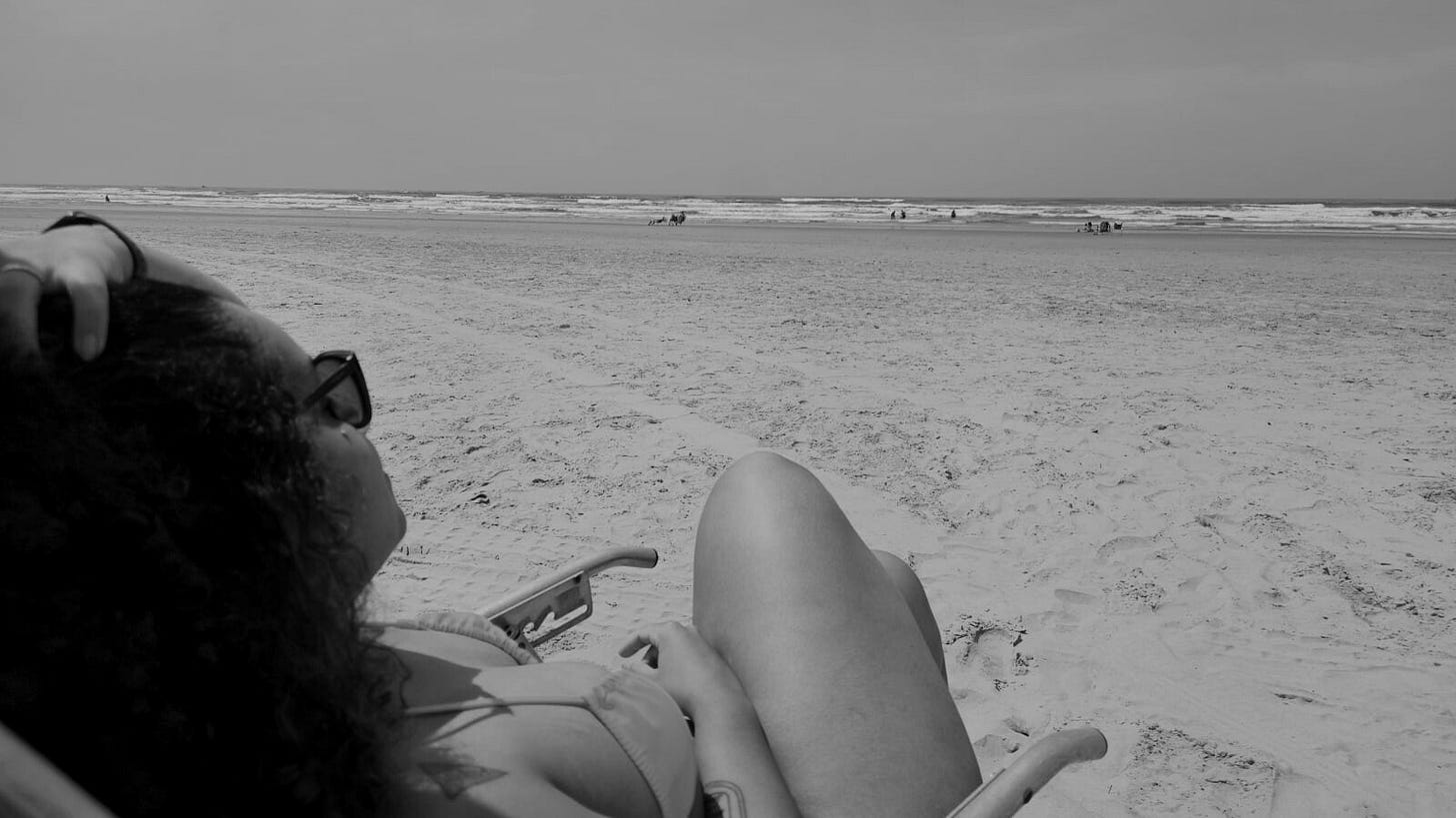 Foto em preto e branco em que apareço sentada numa cadeira olhando o mar e segurando o cabelo