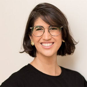 Mulher branca com cabelo escuro, liso e curto usando óculos de aro fino e sorrindo em direção à câmera. Mulher branca com cabelo escuro, liso e curto usando óculos de aro fino e sorrindo em direção à câmera.
