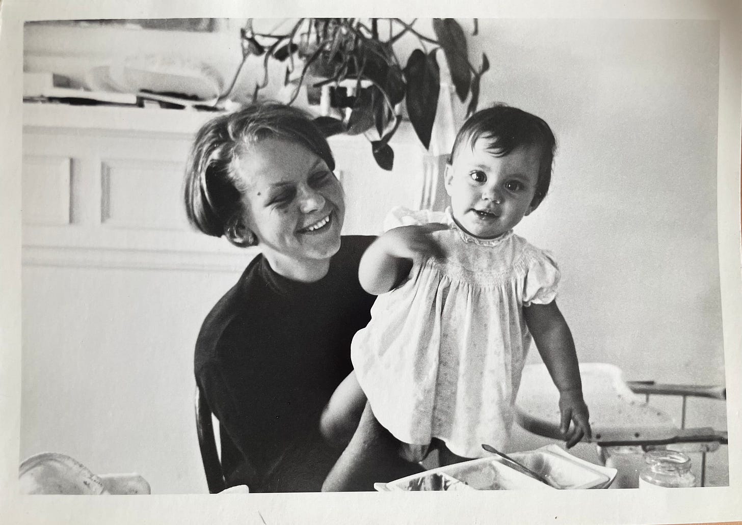 A woman smiles holding a baby to stand on her lap. The baby wears a dress and looks at the camera, smiling. A dish of baby food and a spoon are on the table in the foreground.