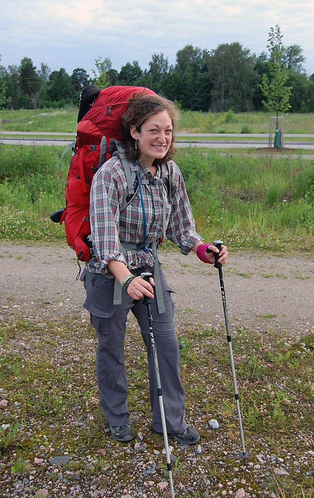 Jenny smiles in front of a field. She's wearing a plaid shirt and a big red backpack, and holding trekking poles.