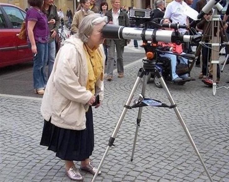 This may contain: an older woman looking through a telescope at people standing on the street and talking to each other