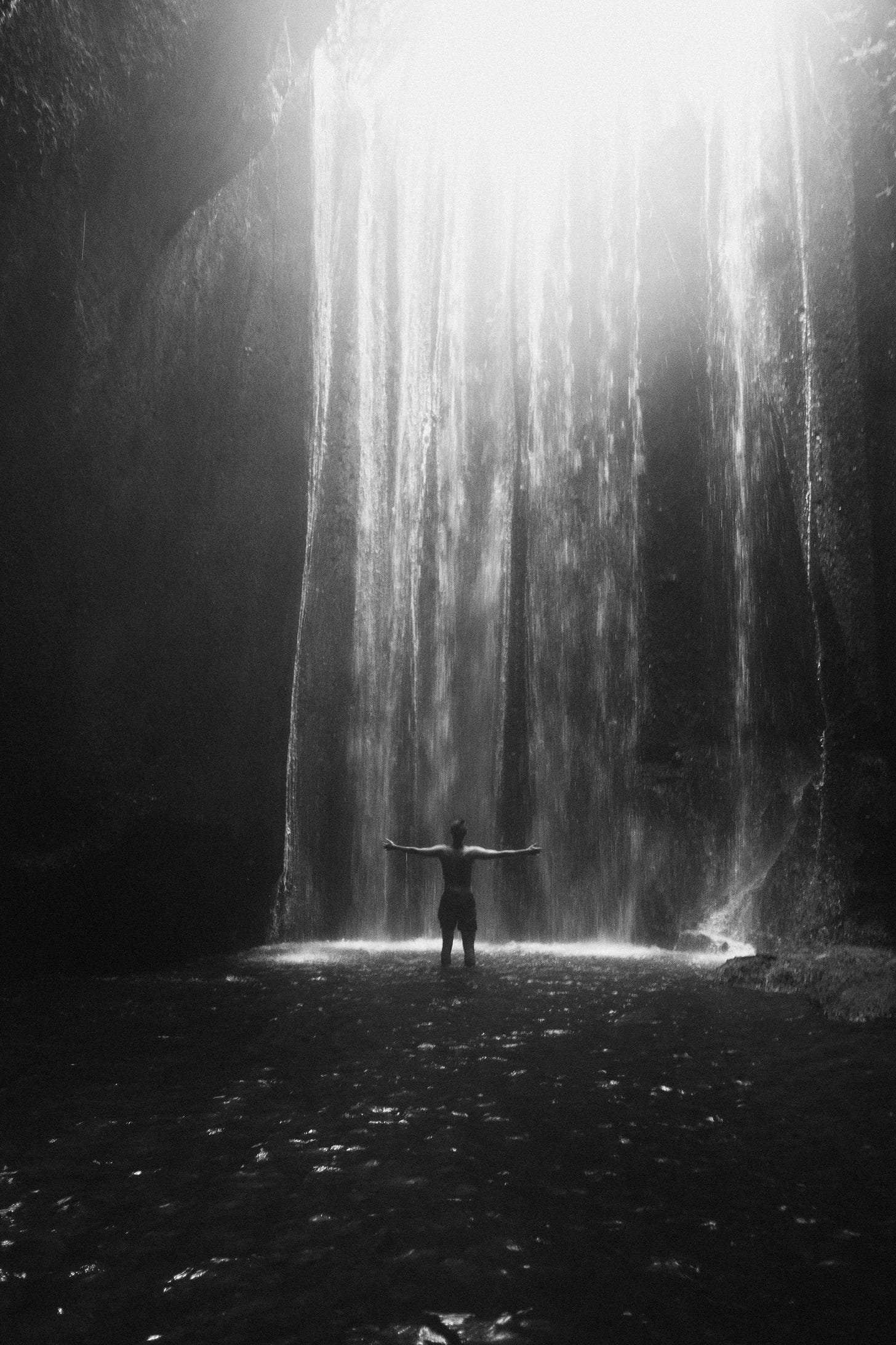 Bright sunbeams piercing through a canyon roof, illuminating a hidden waterfall inside a shadowy, cave-enclosed rock formation.