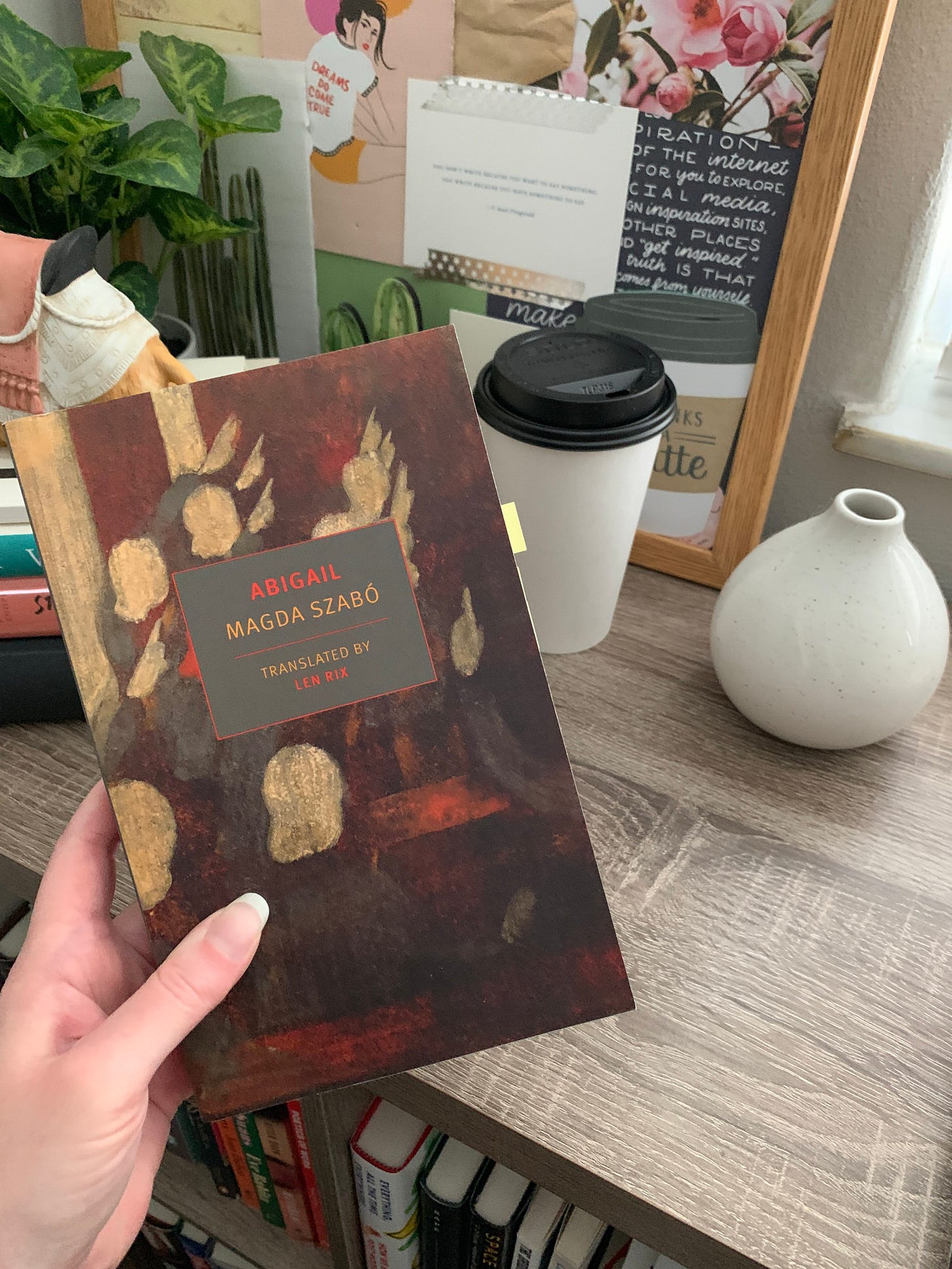hand holding up a copy of Abigail by Magda Szabo, in front of a bookshelf with a moodboard and a cup of coffee