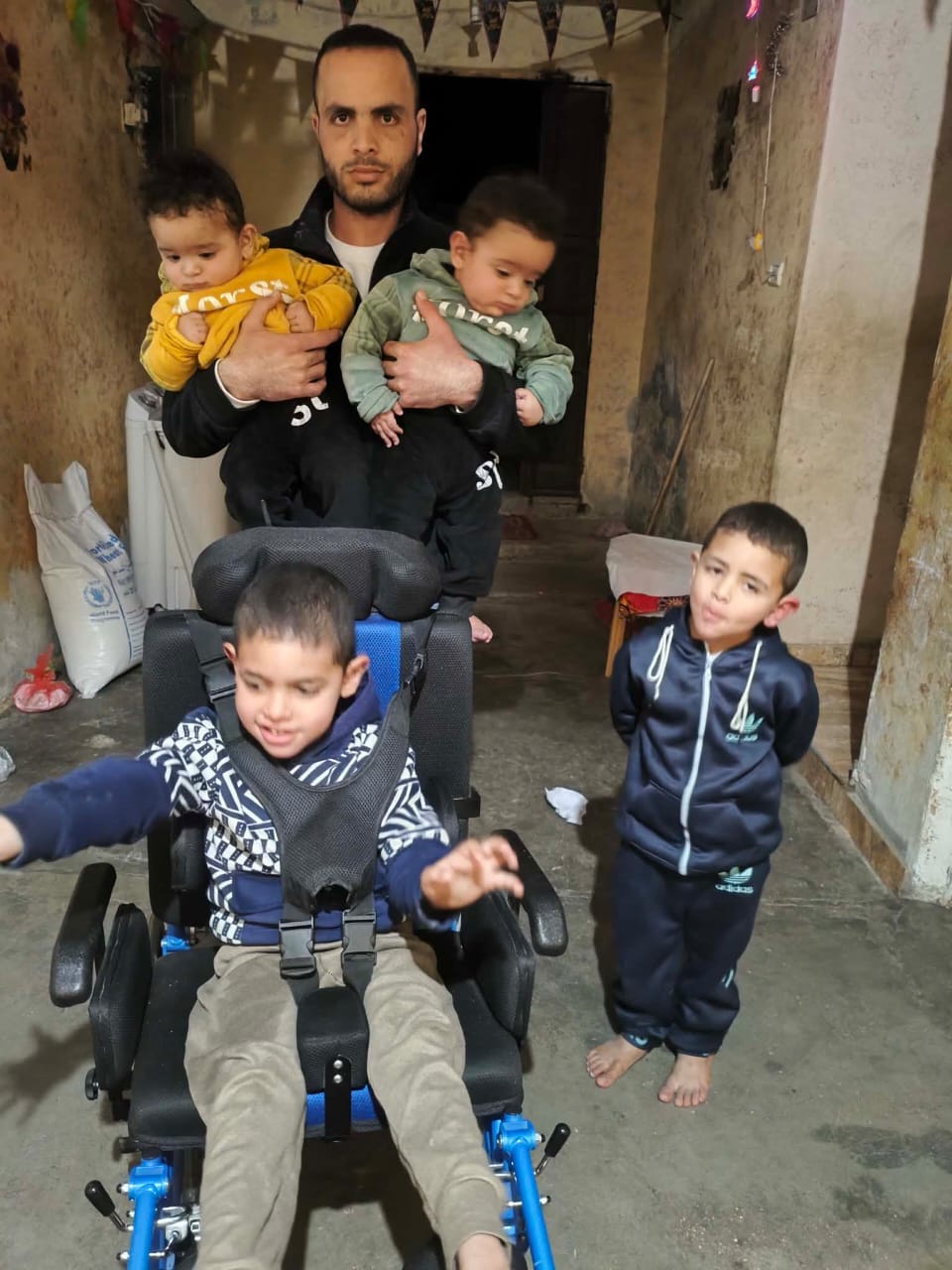 A man stands in a narrow, worn hallway holding two babies, one in each arm. In front of him, a young child sits in a supportive wheelchair with a harness, reaching forward with both hands. Another small child stands barefoot to the right, wearing a dark tracksuit and looking toward the camera. The hallway has rough, aged walls, a concrete floor, and simple household items like sacks and bags placed along the sides. Decorations hang overhead near the entrance behind them. A man stands in a narrow, worn hallway holding two babies, one in each arm. In front of him, a young child sits in a supportive wheelchair with a harness, reaching forward with both hands. Another small child stands barefoot to the right, wearing a dark tracksuit and looking toward the camera. The hallway has rough, aged walls, a concrete floor, and simple household items like sacks and bags placed along the sides. Decorations hang overhead near the entrance behind them.