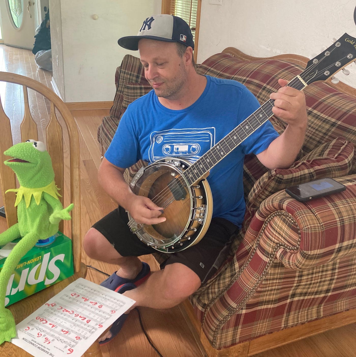 A man in a blue T-shirt holds a banjo and reads sheet music while a puppet of Kermit the Frog sits nearby