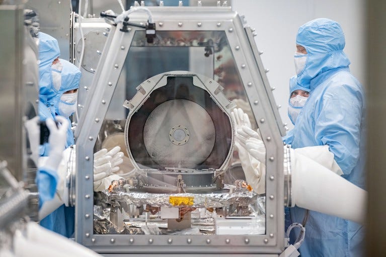 Researchers in PPE in a clean room work with a metal container inside a lab glove box.
